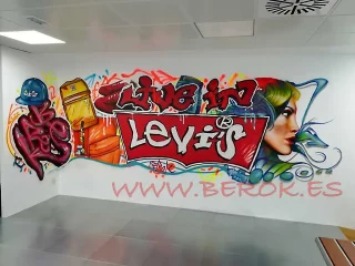 Texto alternativo: Graffiti mural artístico con logo de Levis en oficinas modernas, retrato femenino y objetos urbanos coloridos.