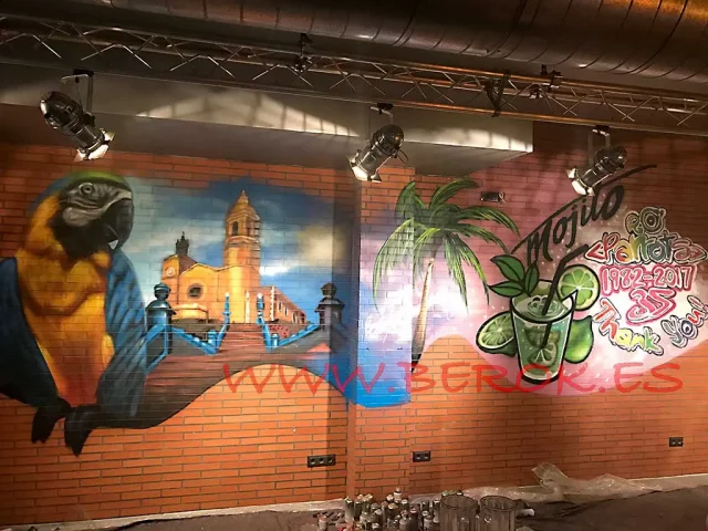 Graffiti realista del Parrots Sitges con guacamayo tropical, palmera e iglesia, pintado en directo por un artista urbano.