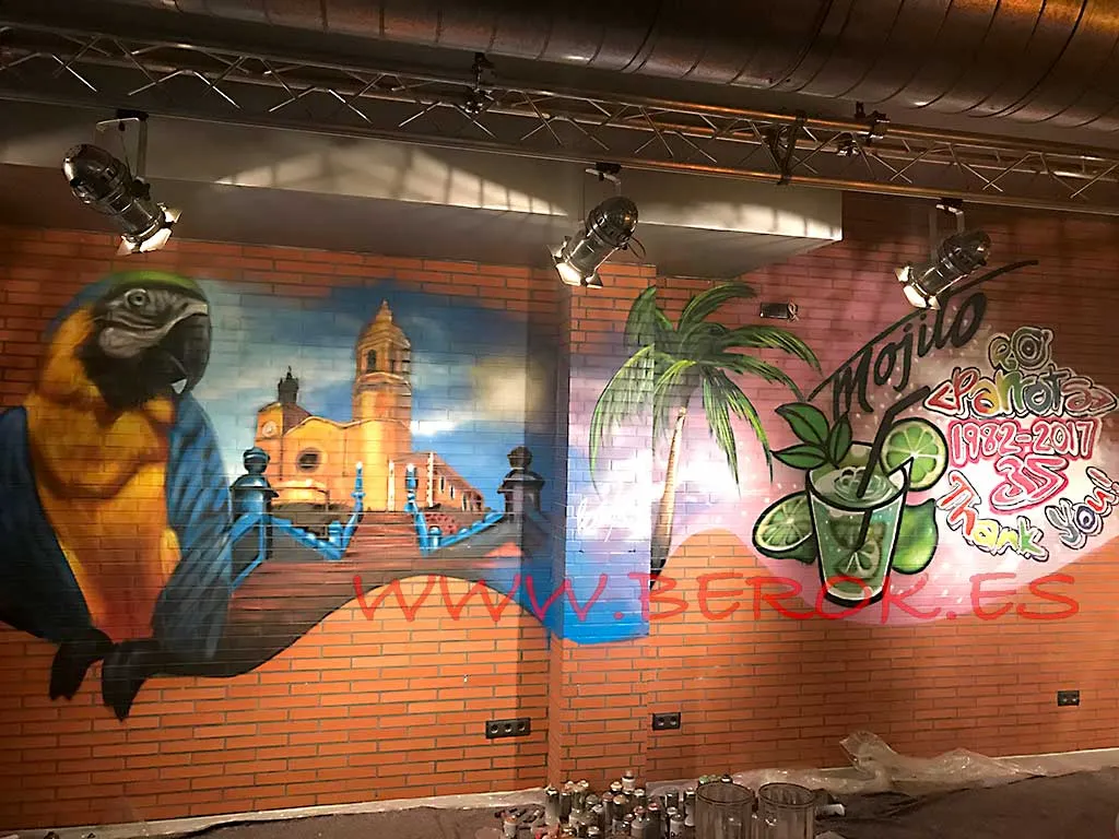 Graffiti realista del Parrots Sitges con guacamayo tropical, palmera e iglesia, pintado en directo por un artista urbano.