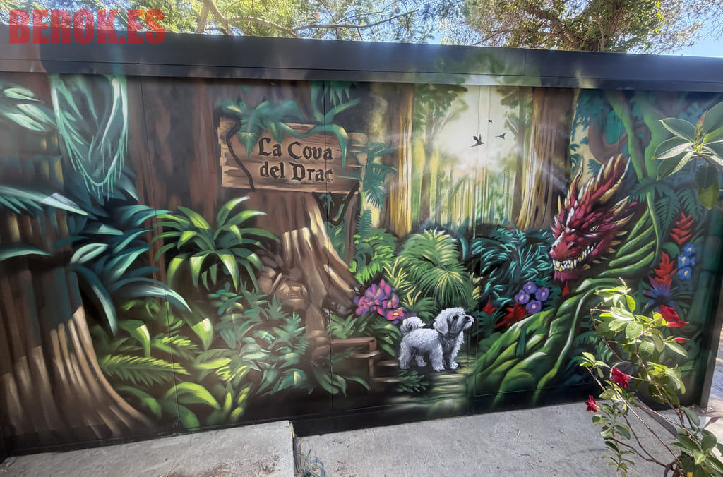 Graffiti de un bosque encantado con un dragón, vegetación tropical, un perro blanco y el rótulo La Cova del Drac pintado sobre una caseta de jardín.