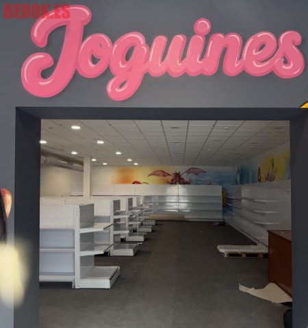 Rótulo "Joguines" en color rosa con efecto volumen sobre la entrada de una tienda de juguetes decorada con murales infantiles.