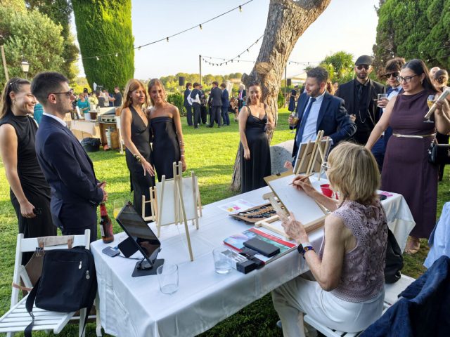 Live art con invitados alrededor de la mesa Invitados reunidos alrededor de una mesa de retratos en acuarela con una artista trabajando en un jardín.