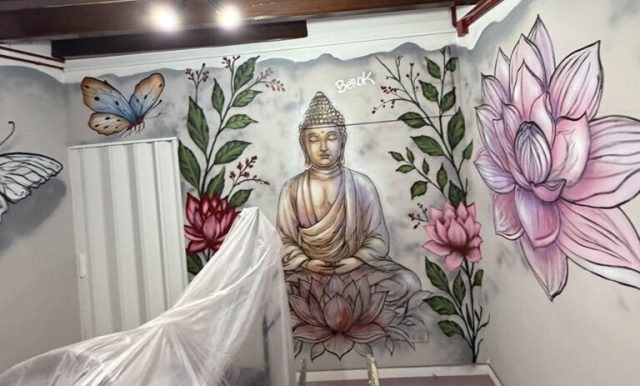 Mural de Buda y loto con escalera de trabajo Vista horizontal del mural de Buda rodeado de hojas y flores de loto, con una escalera delante de la pared.
