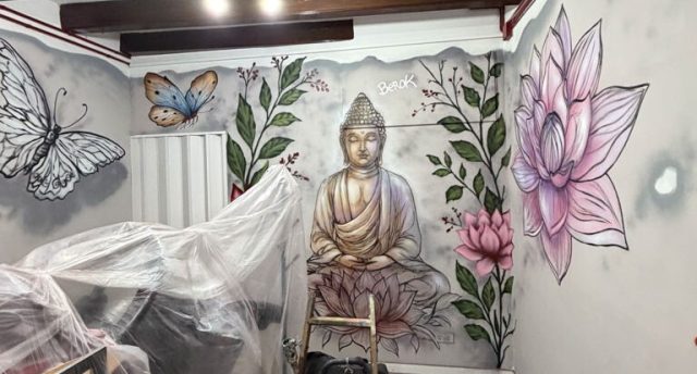 Mural de Buda al fondo de un local Buda entre grandes flores de loto pintado en la pared del fondo de un local con vigas y materiales de trabajo.