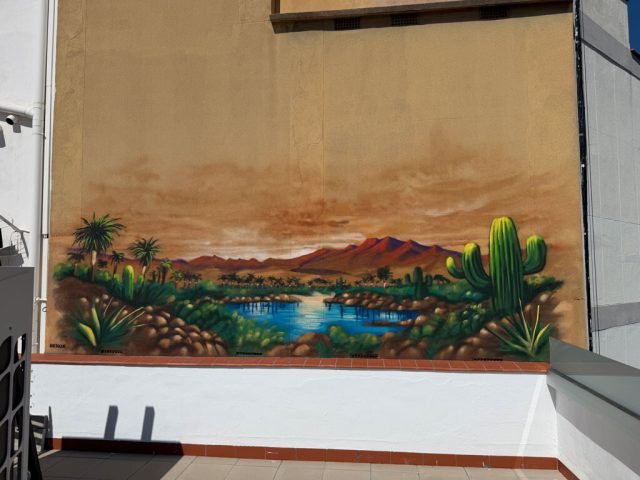 Detalle del lago y los cactus en mural de desierto Detalle de un mural con lago azul, cactus verdes, palmeras y montañas rojizas bajo un cielo cálido.