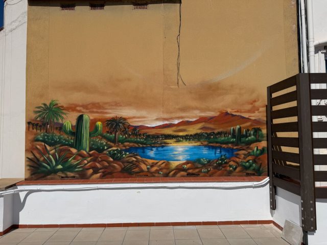 Detalle del mural de lago y cactus junto a celosía Vista cercana del lago azul, las palmeras y los cactus del mural situado junto a una celosía en la terraza.