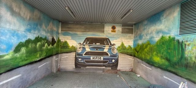Garaje decorado con un coche Mini Cooper de frente al fondo y una carretera con vegetación y cielo en las paredes laterales.