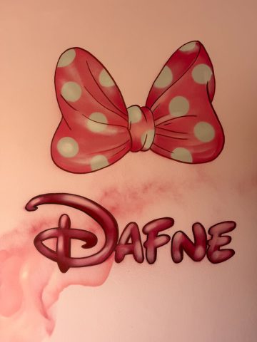 Nombre Dafne pintado en tonos rosas bajo un lazo rojo con lunares sobre una pared rosa.