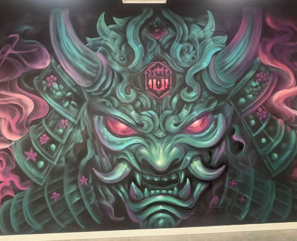 Detalle de una máscara de inspiración samurái con cuernos, dientes y ojos rosas sobre un fondo negro y verde turquesa.