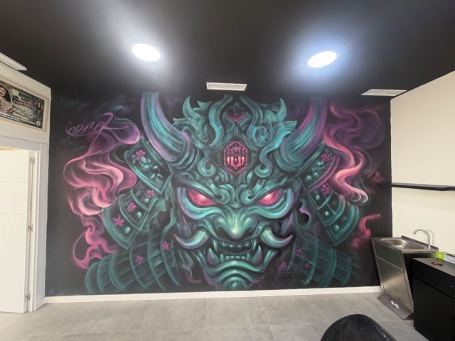 Pared decorada con mural de máscara samurái Vista amplia de una pared interior con una máscara de inspiración samurái en turquesa y rosa sobre fondo oscuro.