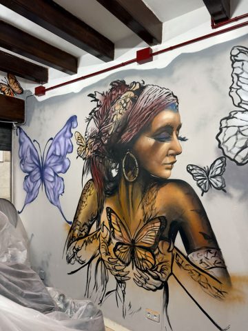 Retrato mural de una mujer de perfil con flores en el cabello y trazos de mariposas sobre el brazo y el hombro.