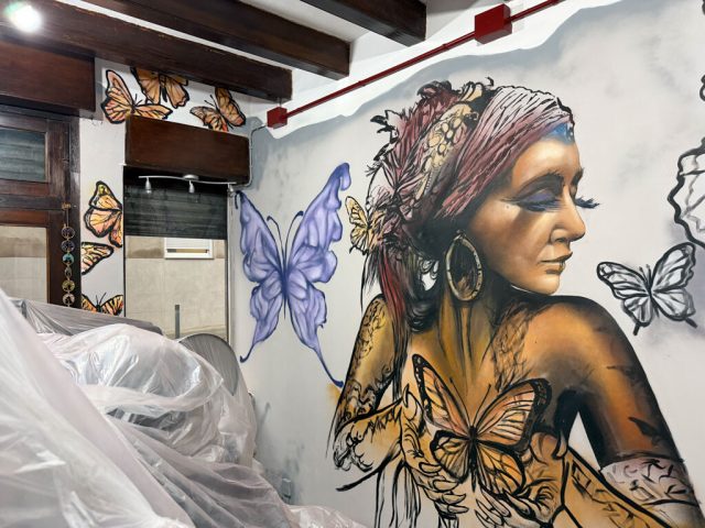 Vista cercana de la mujer de perfil pintada en la pared, con flores en el cabello y una mariposa amarilla en el hombro.