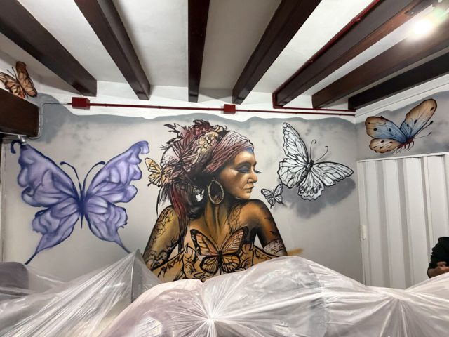Vista horizontal de la pared pintada con una mujer de perfil, una mariposa azul grande y varias mariposas pequeñas.