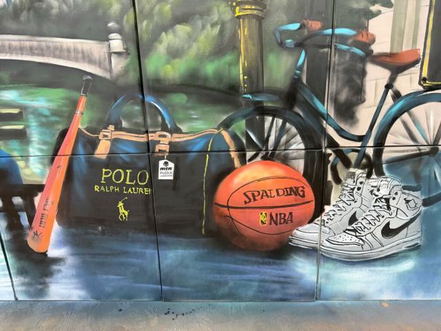 Detalle de un mural de Nueva York con bicicleta, balón de baloncesto, zapatilla y bolsa pintados junto a un paisaje de parque.