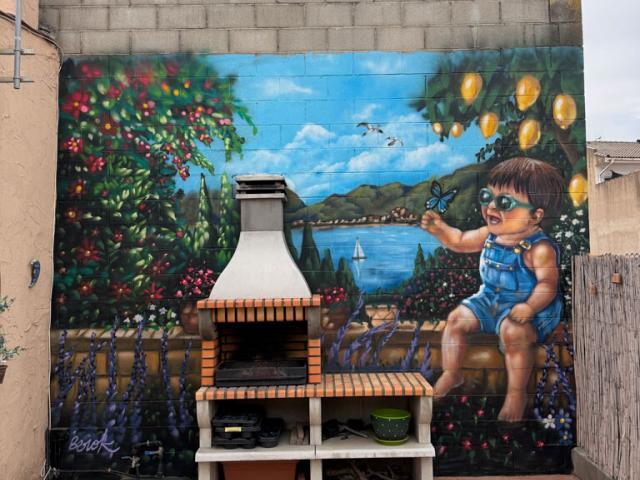 Mural de niña y paisaje en un patio con barbacoa Mural de una niña sentada junto a un paisaje de agua, montañas y vegetación detrás de una barbacoa de obra.