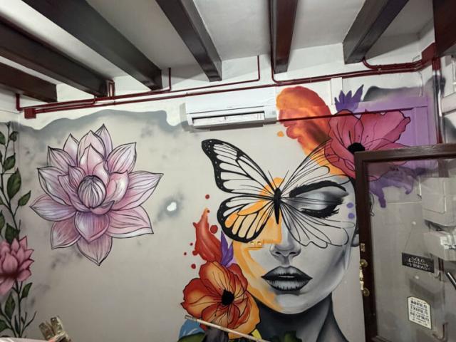 Mural de un rostro femenino en blanco y negro con una gran mariposa amarilla sobre los ojos y flores de colores.