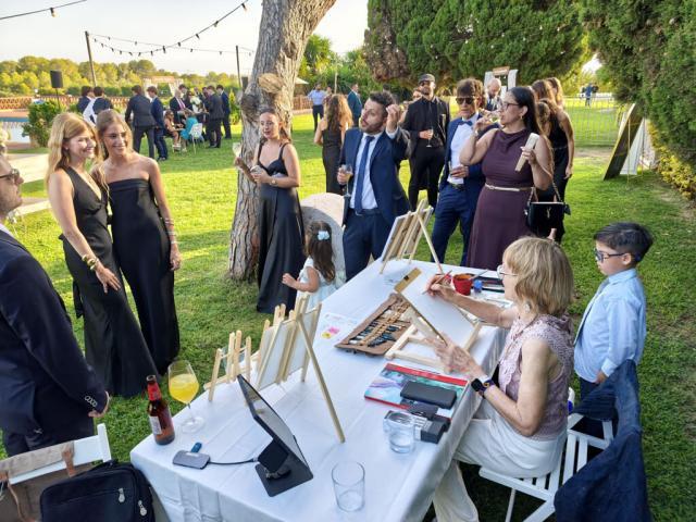 Artista y grupo de invitados en el puesto de retratos Vista desde la mesa de una artista que pinta con acuarelas frente a varios invitados en un jardín con guirnaldas de luces.