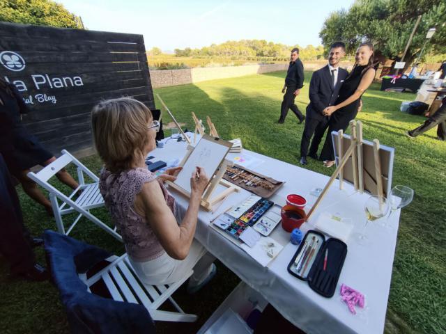 Artista pintando el retrato de una pareja en directo Artista vista de espaldas trabajando con acuarelas mientras una pareja posa delante de ella en un jardín.