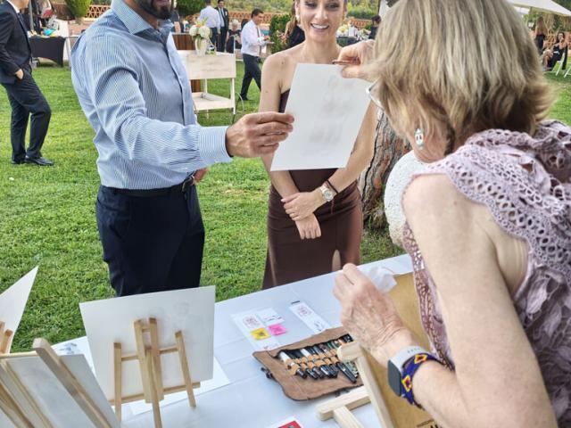 Entrega de una ilustración a una pareja en un evento Un hombre recibe una ilustración de la artista mientras posa junto a una mujer ante la mesa de retratos.