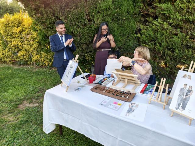 Retratos en directo para una pareja durante un evento Una pareja posa junto a una mesa mientras una artista prepara un retrato en acuarela en un jardín.